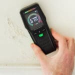 Hand using a moisture meter to measure wall humidity and show how humidity affects indoor paint drying inside a home