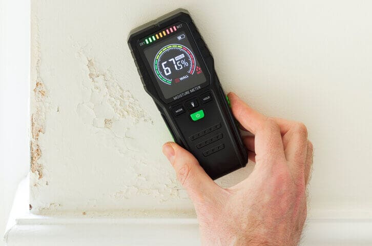 Hand using a moisture meter to measure wall humidity and show how humidity affects indoor paint drying inside a home