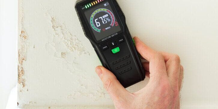 Hand using a moisture meter to measure wall humidity and show how humidity affects indoor paint drying inside a home